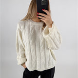 Kvinde iført hvid sweater tager selfie med telefon foran ansigt; langærmet trøje med flettemønster. Hendes negle er malet rød, og hun står mod en neutral baggrund.