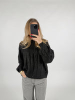 En person tager et billede med en smartphone i et spejlløst rum. De bærer en sort sweater og grå bukser, med hånden i lommen. Baggrunden er enkel og lys.
