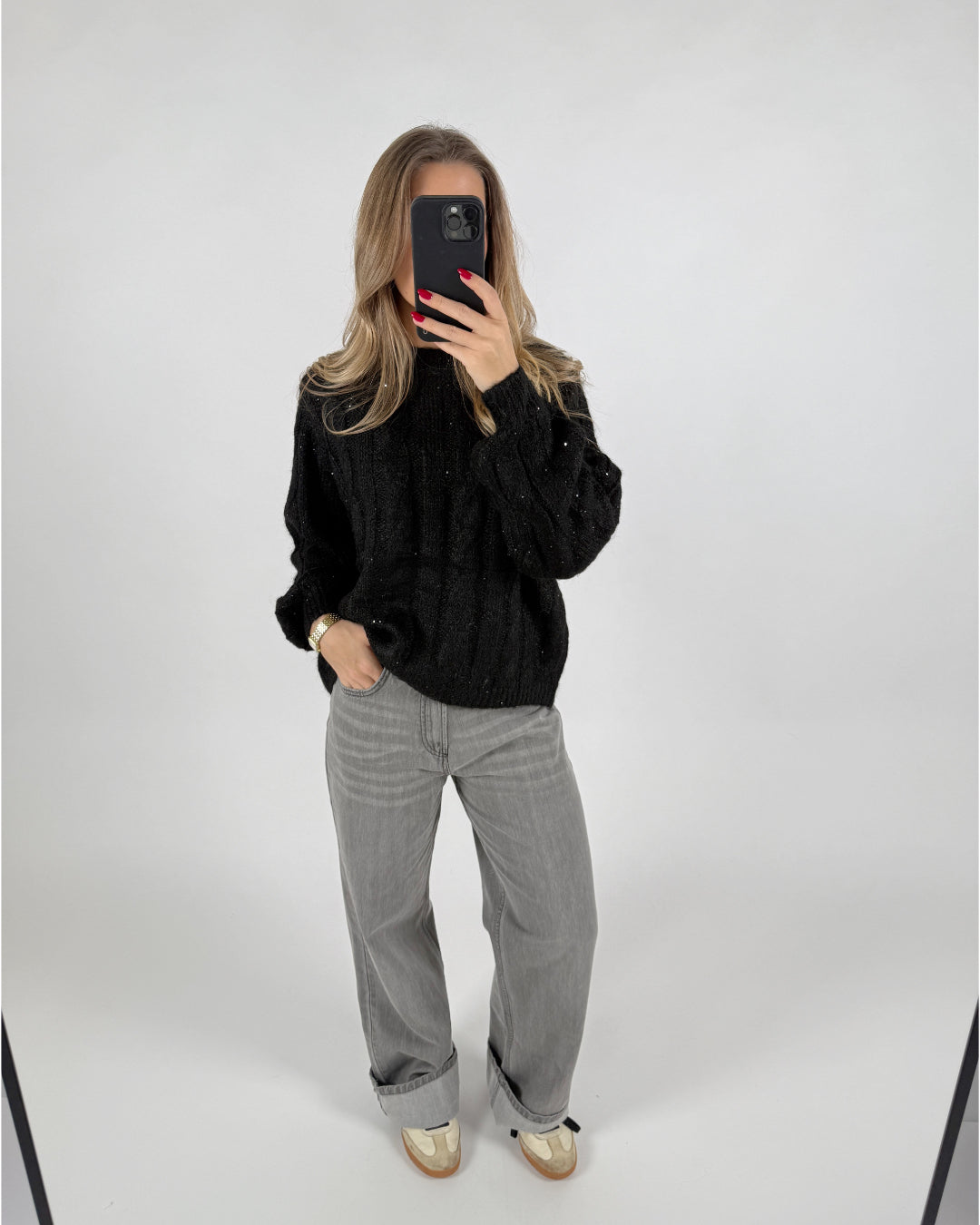 En person står i et minimalistisk rum, iført en sort sweater og grå bukser, mens de tager et selfie med en sort telefon.