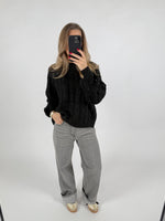 En person står i et minimalistisk rum, iført en sort sweater og grå bukser, mens de tager et selfie med en sort telefon.
