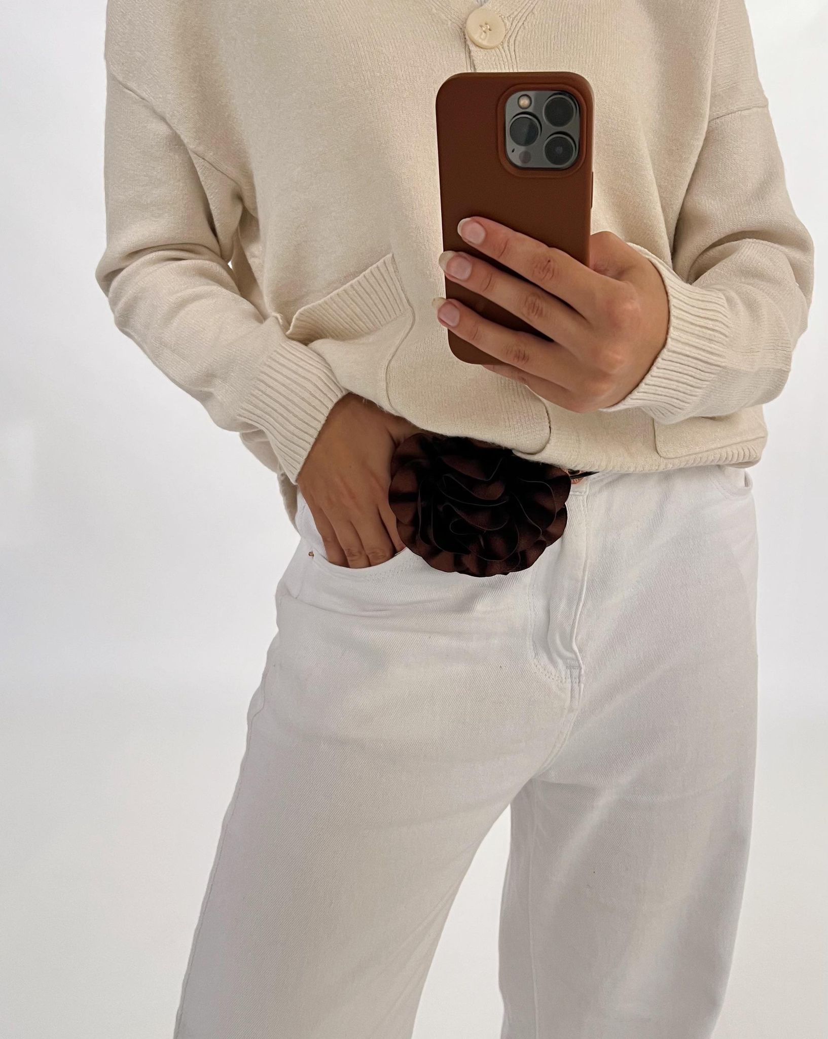 En person holder en telefon og tager et spejlfie iført en lys sweater og hvide bukser, med en brun dekorativ detalje. Baggrunden er hvid og enkel.