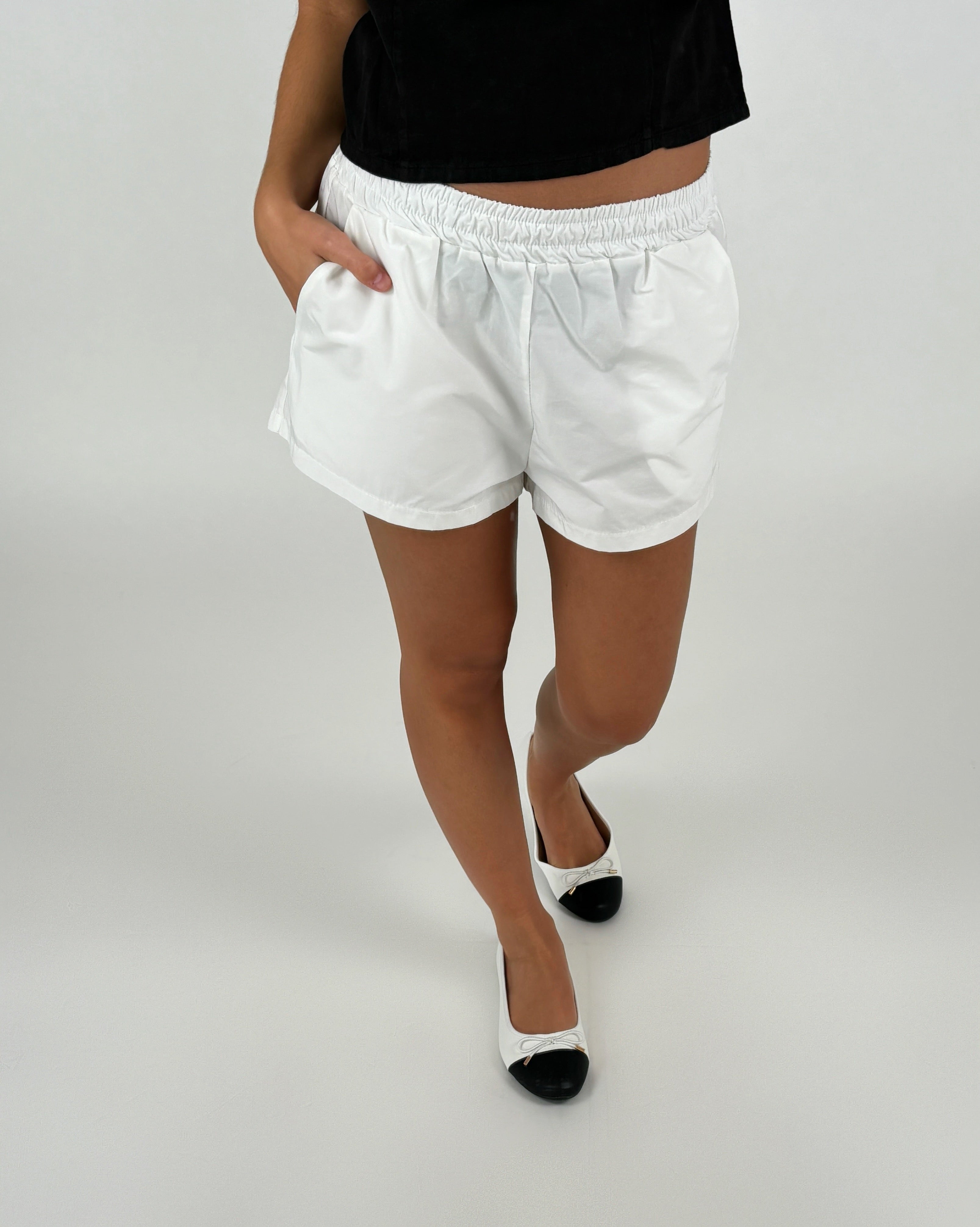 En person iført hvide shorts og sort top står med hænderne i lommerne, iført hvid-sorte sko på et neutralt, lyst gulv.