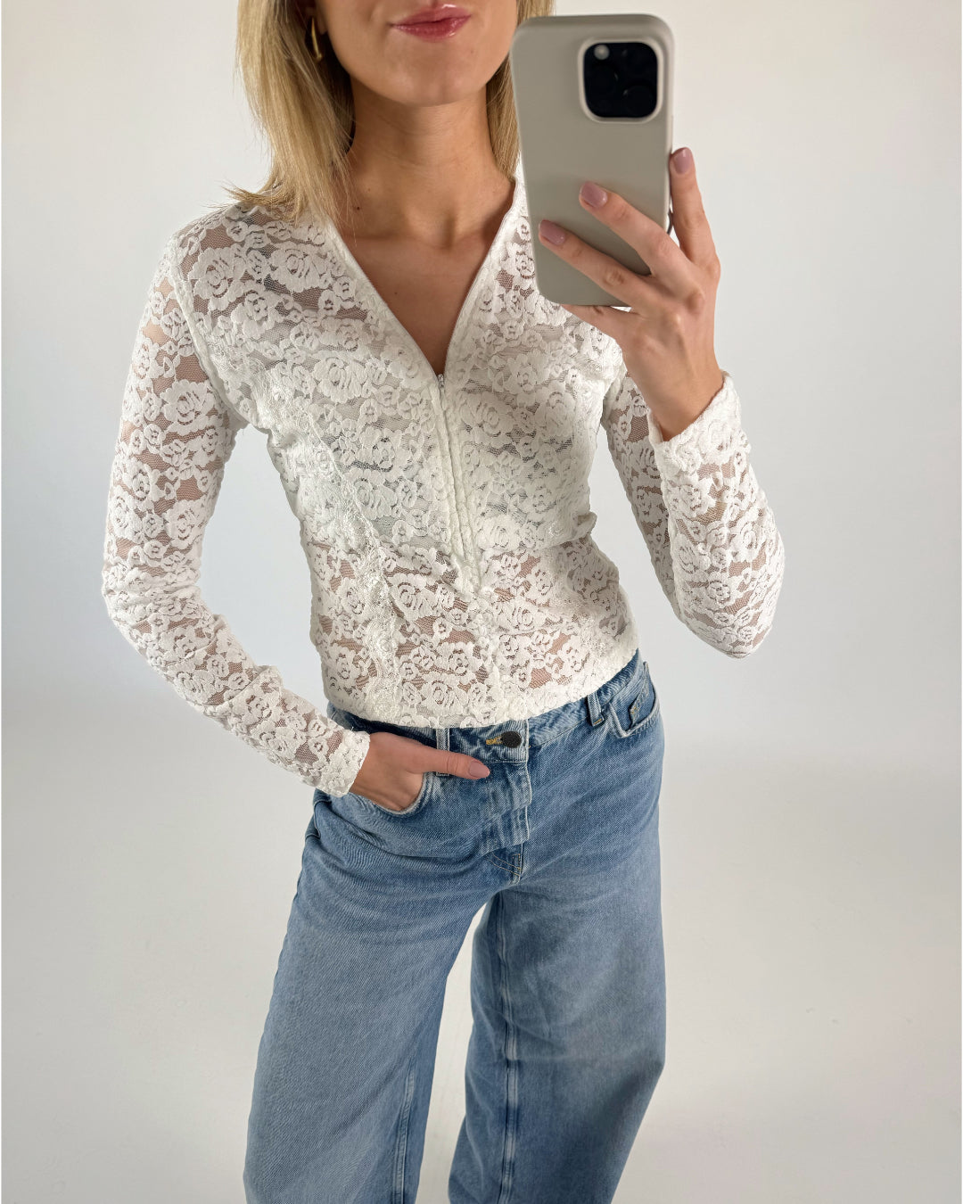 En person iført en hvid blondertrøje og blå jeans tager en selfie med en telefon. Personen står i et lyst, neutralt rum.