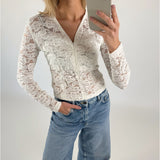 En person iført en hvid blondertrøje og blå jeans tager en selfie med en telefon. Personen står i et lyst, neutralt rum.
