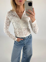 En person iført en hvid blondertrøje og blå jeans tager en selfie med en telefon. Personen står i et lyst, neutralt rum.
