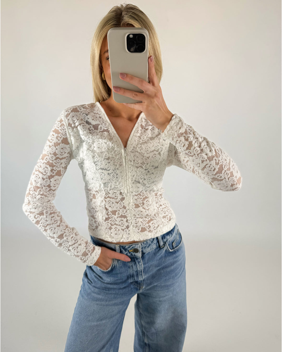 Person tager selfie iført hvid blondetop og blå jeans; står indendørs med hvid væg som baggrund.