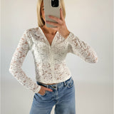 Person tager selfie iført hvid blondetop og blå jeans; står indendørs med hvid væg som baggrund.