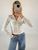 Person tager selfie iført hvid blondetop og blå jeans; står indendørs med hvid væg som baggrund.