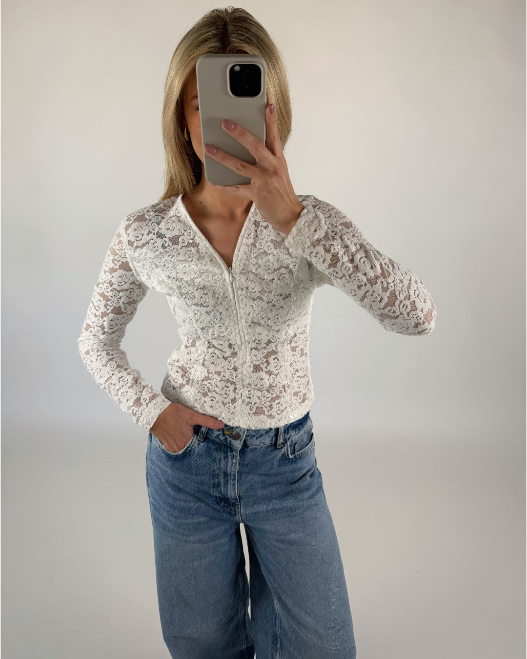 Person tager selfie iført hvid blondebluse og blå jeans; står med en hånd i lommen i et neutralt oplyst rum.