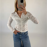 Person tager selfie iført hvid blondebluse og blå jeans; står med en hånd i lommen i et neutralt oplyst rum.
