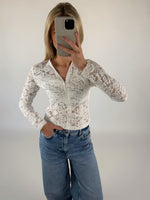 Person tager selfie iført hvid blondebluse og blå jeans; står med en hånd i lommen i et neutralt oplyst rum.