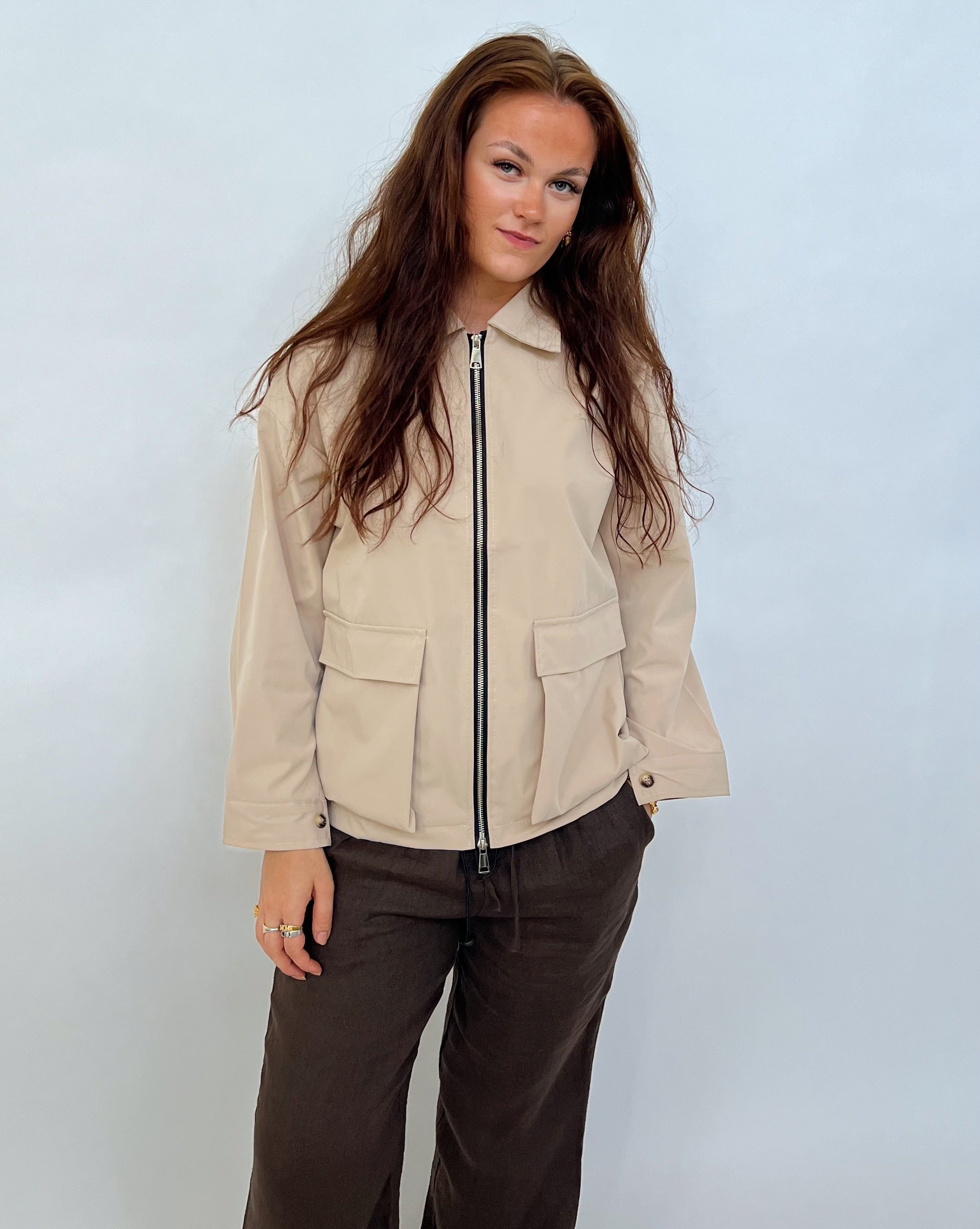 En person med langt hår står afslappet iført en beige jakke med lynlås og brune bukser mod en neutral, lys baggrund.