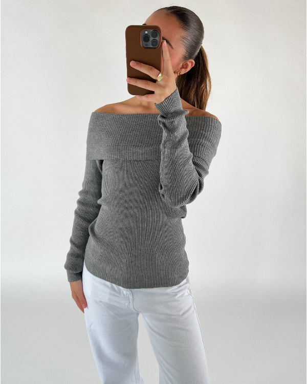 Person tager selfie iført grå off-shoulder trøje og hvide bukser i et neutralt rum. Trøjen har ribstrik, og personen har langt hår i hestehale, ser ind i telefonen.