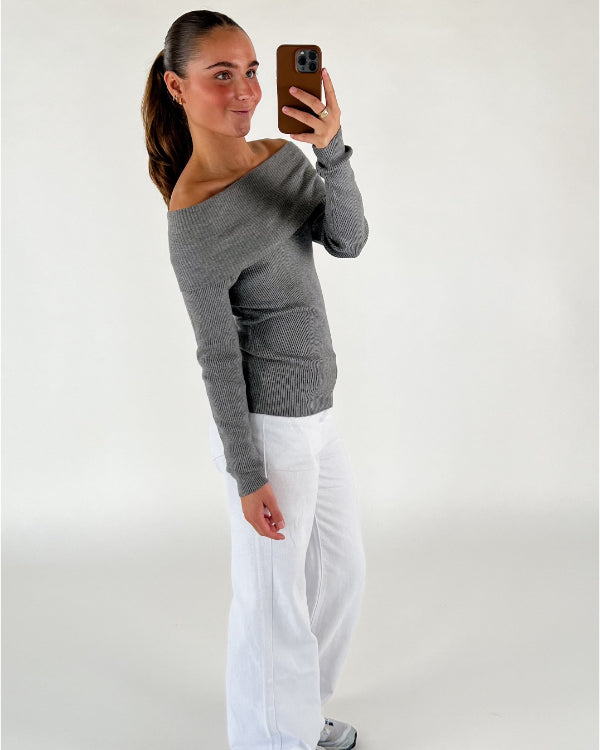 En person tager en selfie i et spejl. Personen bærer en grå off-shoulder sweater og hvide bukser. Baggrunden er en ensfarvet væg.