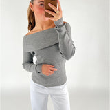 En person iført en off-shoulder grå sweater tager et spejlfoto med en smartphone i brun taske; personen står i et rum med hvid baggrund og bærer hvide bukser.