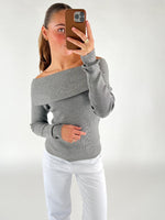 En person iført en off-shoulder grå sweater tager et spejlfoto med en smartphone i brun taske; personen står i et rum med hvid baggrund og bærer hvide bukser.