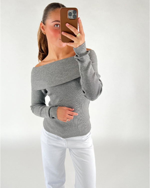 En person iført en off-shoulder grå sweater tager et spejlfoto med en smartphone i brun taske; personen står i et rum med hvid baggrund og bærer hvide bukser.