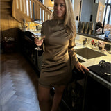 En kvinde i en beige kjole holder en cocktail ved en bar. Baggrunden indeholder en trappe og barudstyr, mens lyset skaber en hyggelig atmosfære.