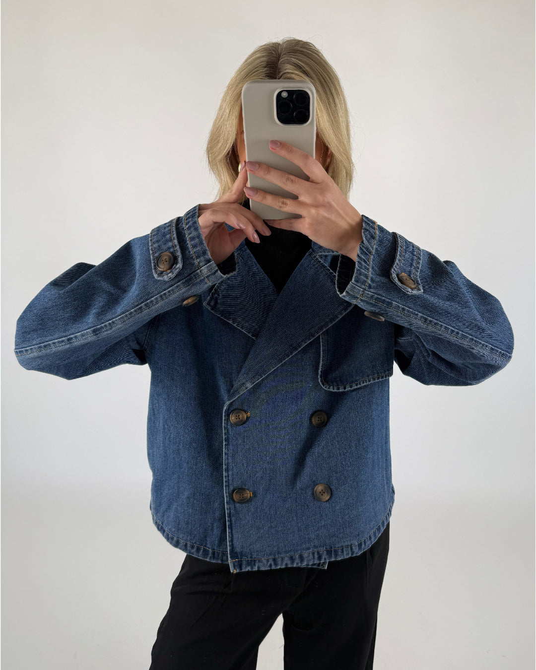 En person i en dobbeltknappet denimjakke tager et spejlselfie med en smartphone i lyse omgivelser. Personen har sort tøj under jakken.