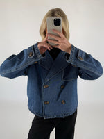En person i en dobbeltknappet denimjakke tager et spejlselfie med en smartphone i lyse omgivelser. Personen har sort tøj under jakken.