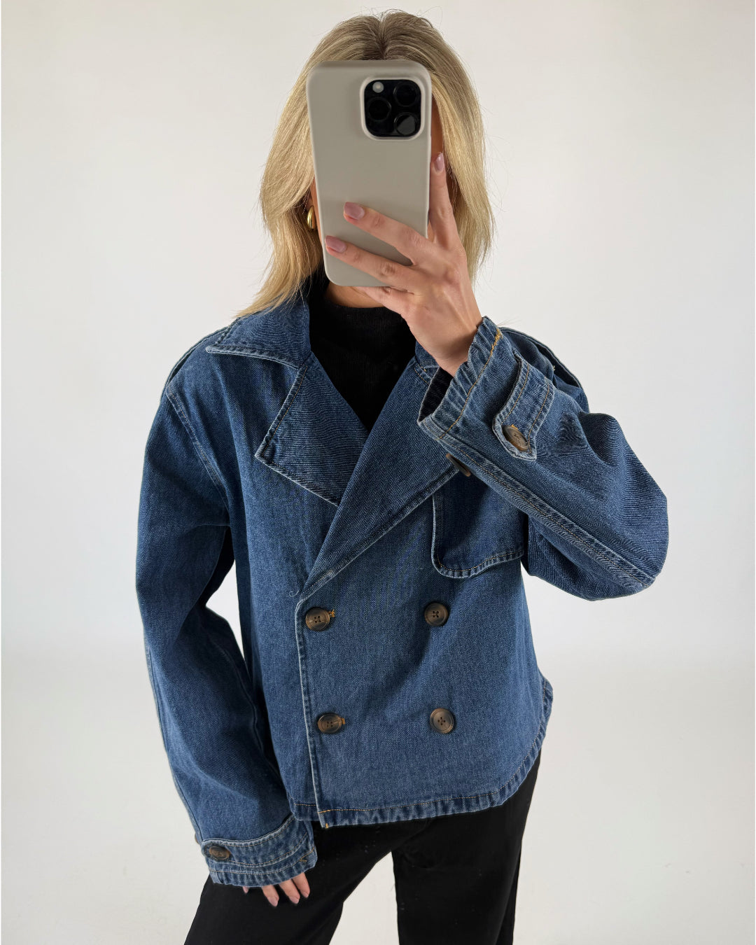 Person i blå denimjakke tager et spejlbillede med en smartphone, står i et rum med en hvid baggrund.