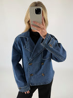 Person i blå denimjakke tager et spejlbillede med en smartphone, står i et rum med en hvid baggrund.