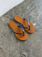 Et par brune sandaler med blå stropper ligger stille på en ru, grå cementoverflade ved siden af en hvid væg.