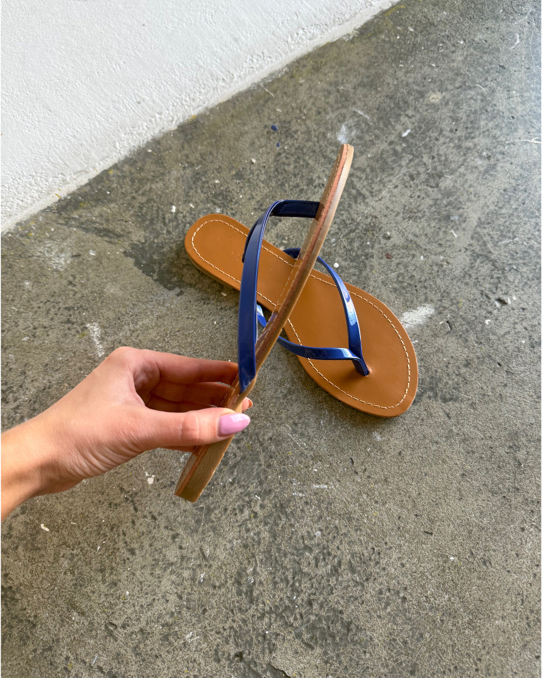 En hånd holder en brun sandal med blå stropper, mens den ligger på en grå betonoverflade ved siden af en hvid væg.