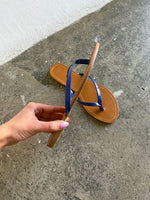 En hånd holder en brun sandal med blå stropper, mens den ligger på en grå betonoverflade ved siden af en hvid væg.