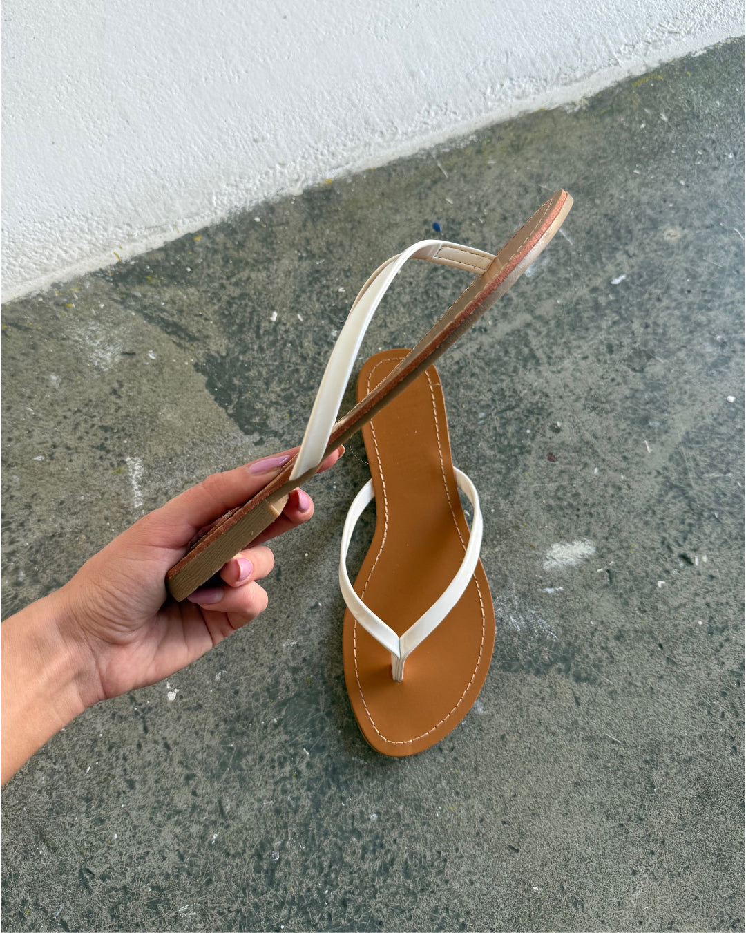 En hånd holder en brun og hvid flip-flop-sandal. Baggrunden er et råt, gråt betonunderlag ved en hvid væg.