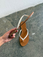 En hånd holder en brun og hvid flip-flop-sandal. Baggrunden er et råt, gråt betonunderlag ved en hvid væg.