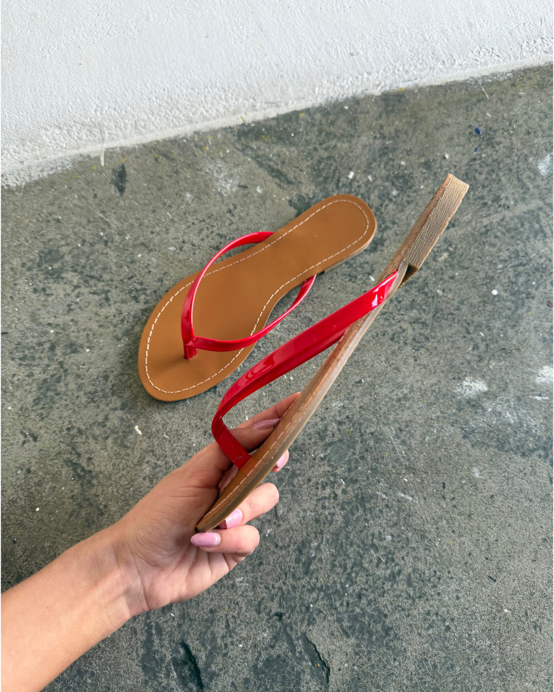 En flip-flop med røde remme holdes i en hånd mod en ru, grå cementbaggrund nær en hvid væg. En anden lignende sandal ligger på gulvet.