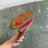 En flip-flop med røde remme holdes i en hånd mod en ru, grå cementbaggrund nær en hvid væg. En anden lignende sandal ligger på gulvet.