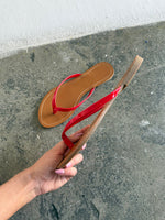 En flip-flop med røde remme holdes i en hånd mod en ru, grå cementbaggrund nær en hvid væg. En anden lignende sandal ligger på gulvet.