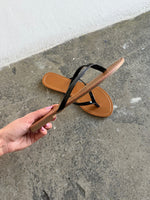 En brun og sort flip-flop sandal holdes i hånden over en grov betonoverflade, mens en anden sandal ligger på gulvet ved siden af en hvid væg.