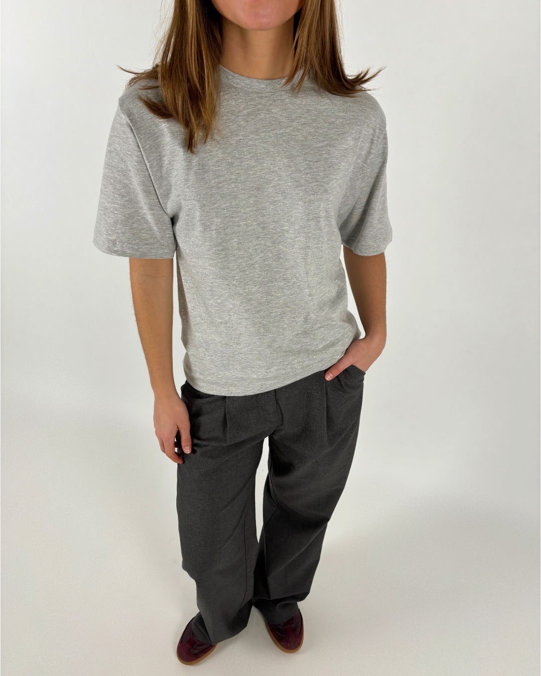 En person bærer en grå t-shirt og mørke bukser, står med hænderne i lommerne. Baggrunden er neutral hvid, hvilket giver et minimalistisk look.