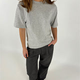 En person bærer en grå t-shirt og mørke bukser, står med hænderne i lommerne. Baggrunden er neutral hvid, hvilket giver et minimalistisk look.