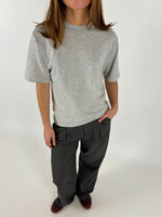 En person bærer en grå t-shirt og mørke bukser, står med hænderne i lommerne. Baggrunden er neutral hvid, hvilket giver et minimalistisk look.