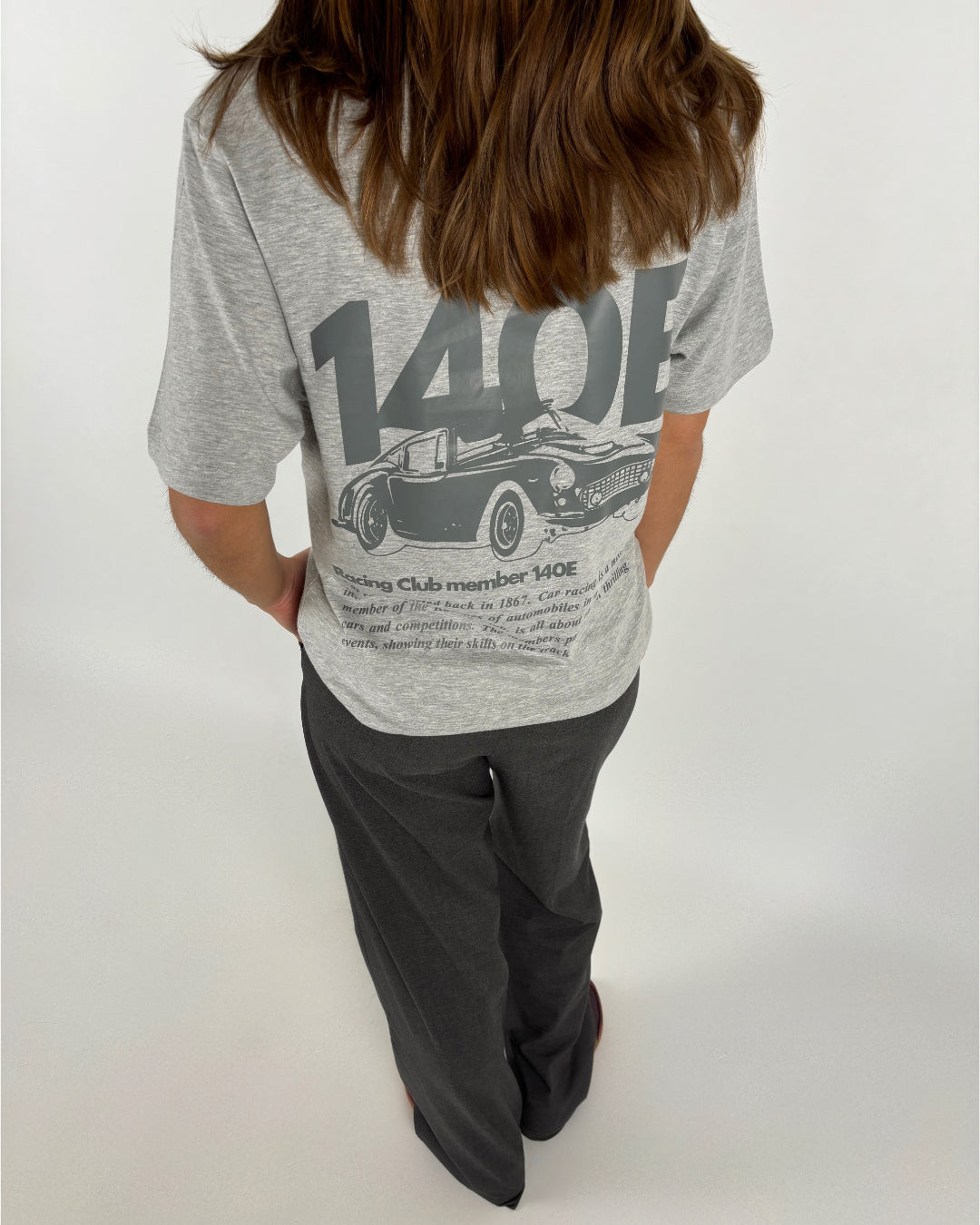 En person iført grå T-shirt med sort-hvidt billede og tekst om racerbiler står med ryggen til, i et neutralt, lyst rum. Tekst på T-shirten: "140E", "Racing Club member 140E," detaljer om bilklubben.
