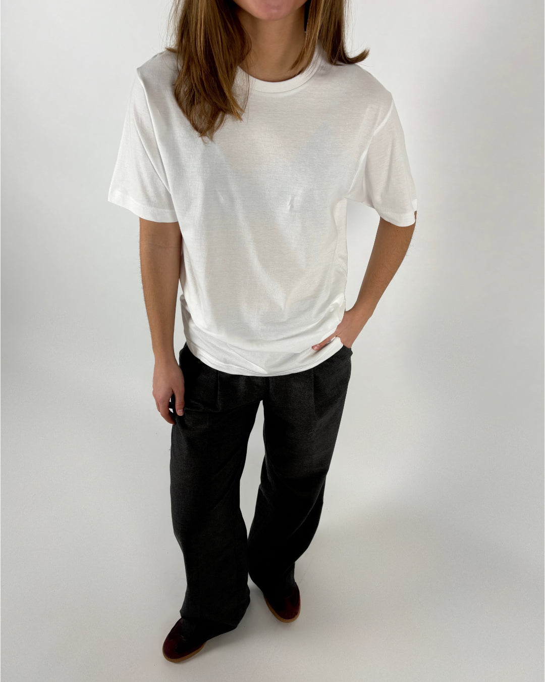 En person iført hvid t-shirt og sorte bukser står med hænderne i lommerne, fotograferet mod en grå baggrund.