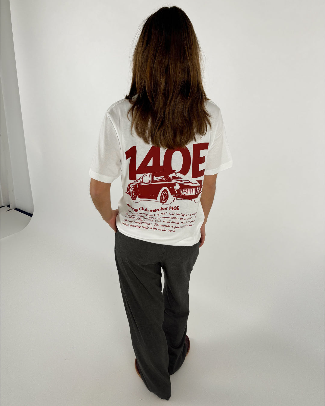 En person står med ryggen til iført en hvid T-shirt med rød grafik af en bil og tekst "140E Racing Club member 140E ... 1987 ... Car racing ...". De er i en lys indendørs omgivelser.