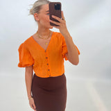 Person tager en selfie iført en orange bluse og brun nederdel, med højt opsat hår, mod en enkel, lys baggrund. Personen holder en mobiltelefon, der dækker ansigtet.