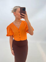 Person tager en selfie iført en orange bluse og brun nederdel, med højt opsat hår, mod en enkel, lys baggrund. Personen holder en mobiltelefon, der dækker ansigtet.