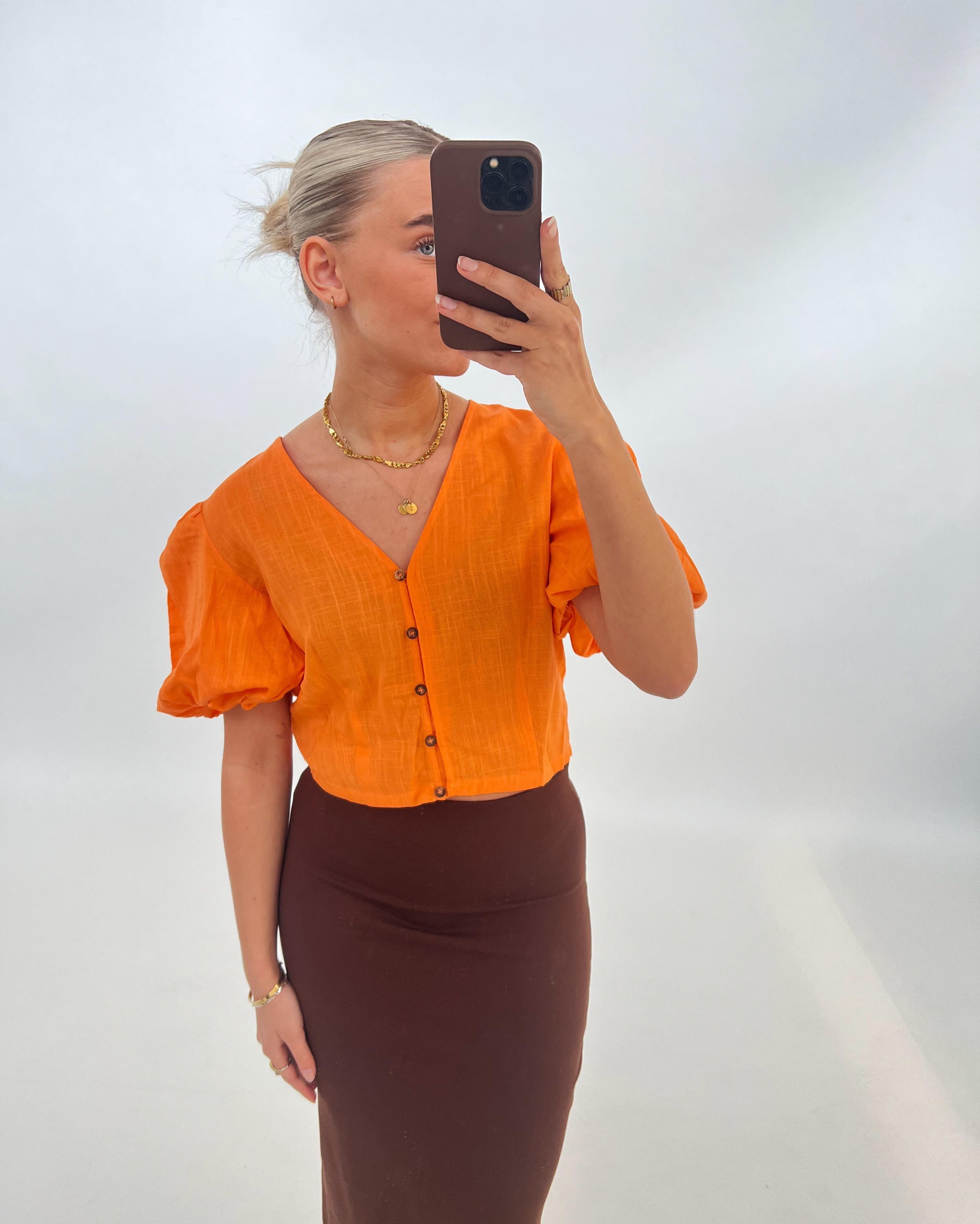 Person tager en selfie iført en orange bluse og brun nederdel, med højt opsat hår, mod en enkel, lys baggrund. Personen holder en mobiltelefon, der dækker ansigtet.