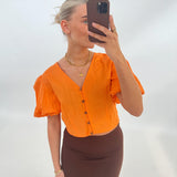 En person tager en selfie iført en orange bluse og brun nederdel, stående foran en lys baggrund. Personen holder en mobiltelefon, og bærer smykker.