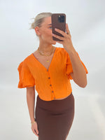 En person tager en selfie iført en orange bluse og brun nederdel, stående foran en lys baggrund. Personen holder en mobiltelefon, og bærer smykker.