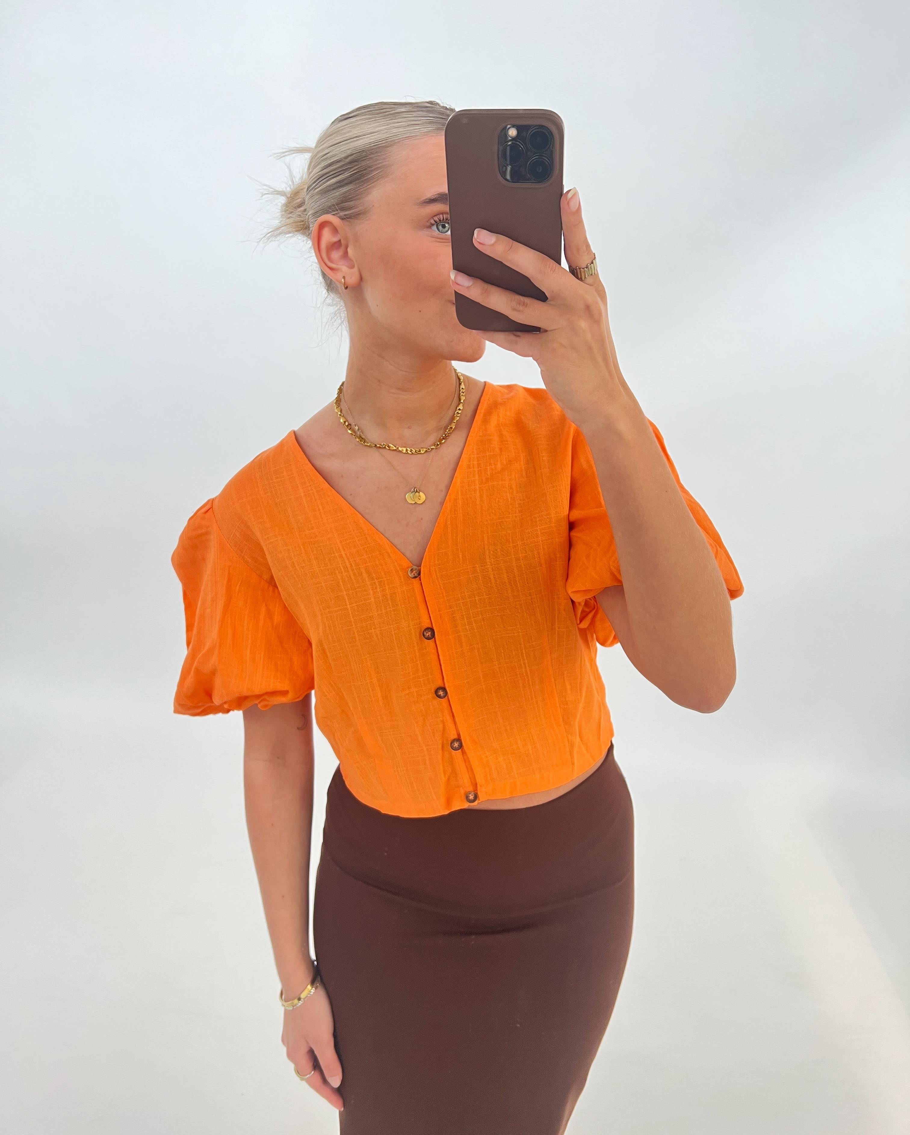En person tager en selfie iført en orange bluse og brun nederdel, stående foran en lys baggrund. Personen holder en mobiltelefon, og bærer smykker.