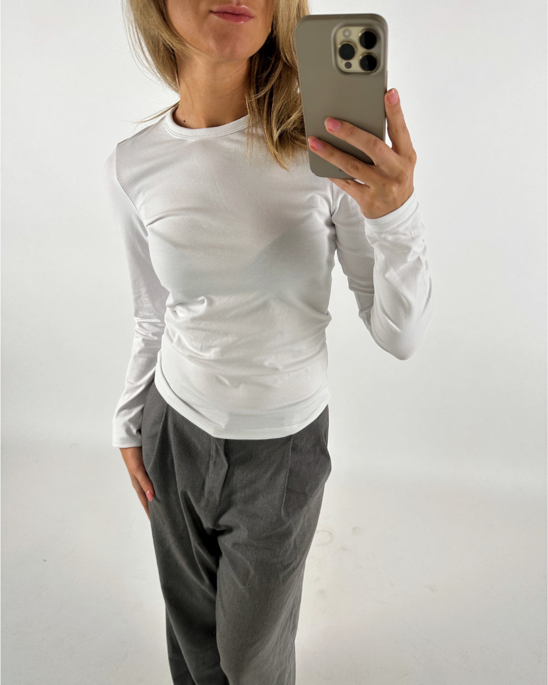 En person iført en hvid trøje og grå bukser holder en smartphone foran et hvidt baggrund. Personen ser ud til at tage et selfie.