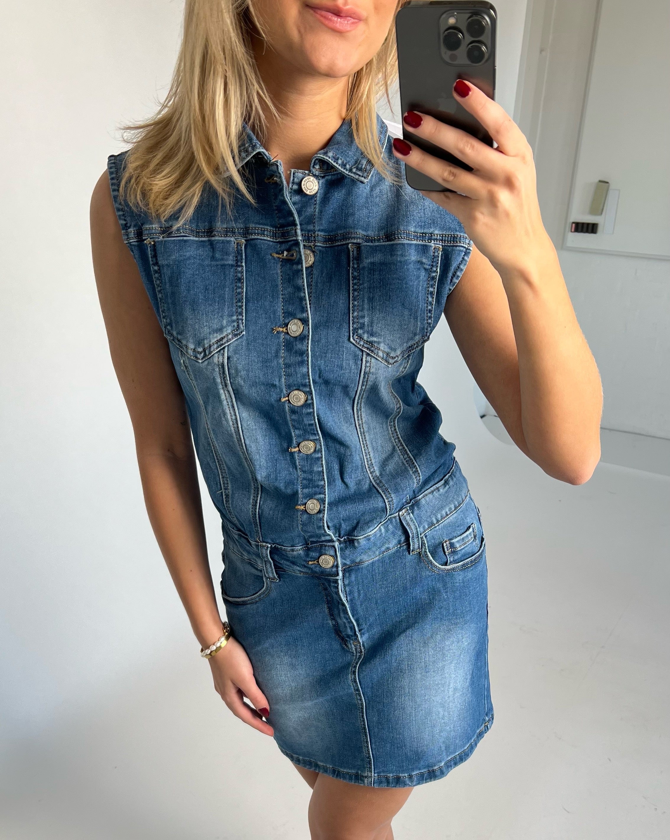 Kvinde i en ærmeløs denimkjole tager et spejlfoto af sig selv. Hun har rødmalede negle og et armbånd. Baggrunden er et lyst rum med simple vægge.