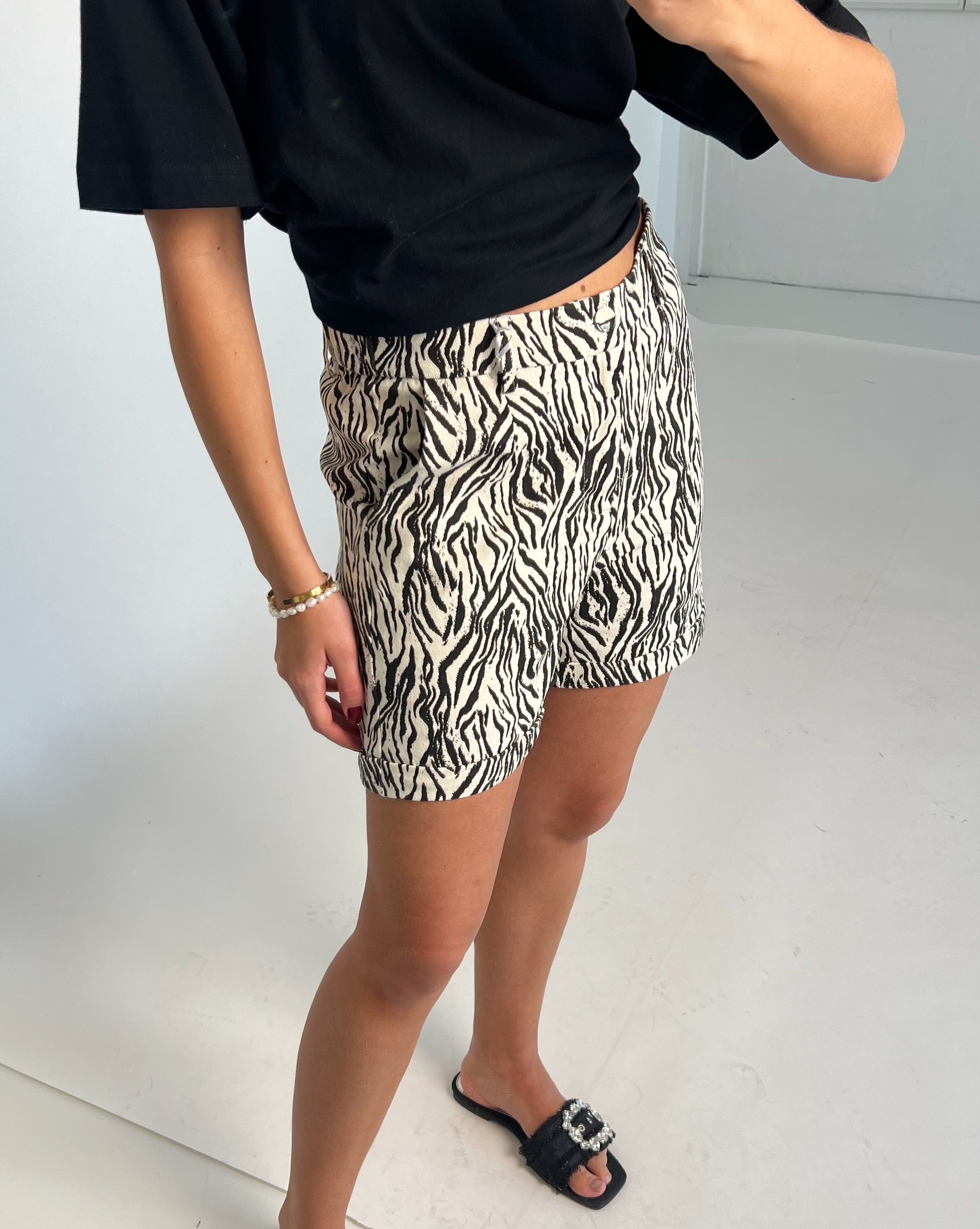 Kvinde i zebramønstrede shorts står i et minimalistisk, lyst rum. Hun bærer en sort top og sorte sandaler med et spænde. Der er ingen synlig tekst.